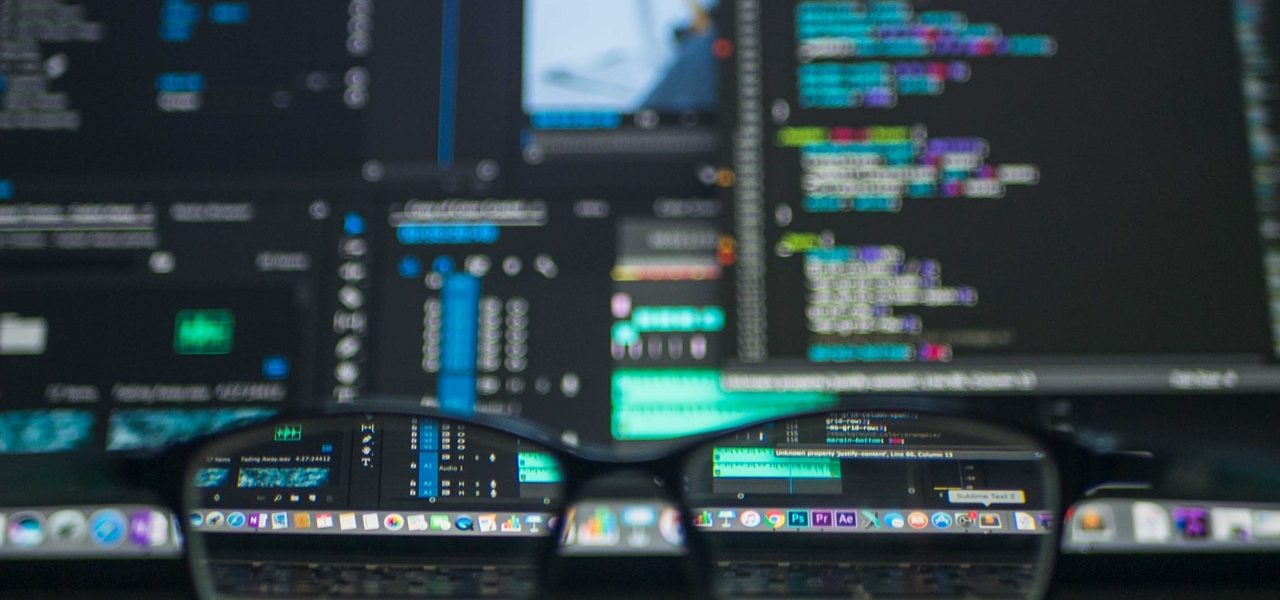 Glasses frame on a desk in front of a computer screen displaying code Glasses frame on a desk in front of a busy computer screen displaying code